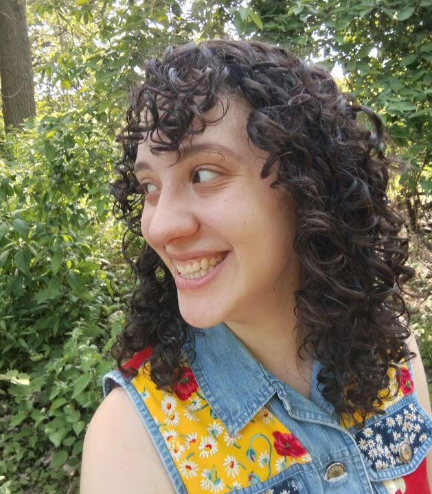Monica headshot A photo of me, Monica! I'm a young light-skinned Latina woman with curly brown hair wearing a patchwork denim vest. I'm standing in front of foliage and laughing.